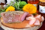 Zdjęcie przedstawia produkt regionalny – schab dojrzewający. Schab z pokrojonymi plasterkami mięsa ułożony jest na drewnianej desce wśród kwiatów i ziół
