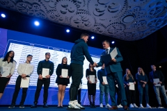Członek Zarządu Zdzisław Szwed wręcza stypendium chłopakowi w wieku szkolnym. Za  nimi stoi kilkunastu laureatów stypendium