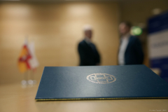 Widok na teczkę z podpisaną umową. W tle - dwie rozmazane postacie oraz niewyraźne, spoczywające flagi.