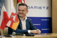 Łukasz Kutyba członek zarządu firmy Adamietz uśmiechający się i spoglądający w swoją prawą stronę. Siedzi przy stole - leży przy nim teczuszka, długopis oraz mikrofon. Na stole widoczne są również dwie małe flagi: Polski oraz Województwa Lubelskiego.