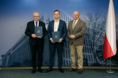 Piotr Matej, Łukasz Kutyba oraz Jarosław Stawiarski pozujący do wspólnego zdjęcia. Dwóch pierwszych trzyma w dłoniach podpisane umowy. Po prawej stronie zdjęcia widoczna jest duża flaga Polski.