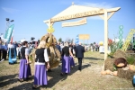 widok na bramę dożynkową, którą przekraczają panie w czarno-fioletowo-białych strojach ludowych, niosąc wieniec dożynkowy. Dookoła w tle widać ludzi obserwujących korowód.