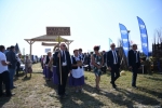 Grupa ludzi w strojach ludowych przekracza bramę dożynkową. Dookoła w tle widać ludzi obserwujących korowód.