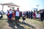 Przez bramę dożynkową przechodzą członkinie koła gospodyń wiejskich w tradycyjnych strojach ludowych. na czele idzie starsza pani i mała dziewczynka. Dookoła w tle widać ludzi obserwujących korowód.