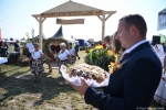 Z prawej strony kadru, poza polem ostrości widać starostę dożynkowego, który trzyma chleb. W tle przez bramę dożynkową przechodzą uczestnicy korowodu niosący wieniec dożynkowy.