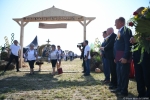 Z prawej strony kadru stoją gospodarze dożynek z marszałkiem województwa na czele. z lewej przez  bramę dożynkową przechodzą uczestnicy korowodu niosący wieniec dożynkowy.