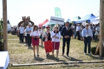 Przez bramę przechodzą reprezentanci Tarnawatki, w tle widać panów niosących duży wieniec. W tle dookoła ludzie obserwujący korowód.