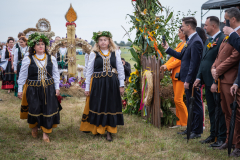Przechodzący przez bramę dożynkową korowód wieńcowy. Dwie kobiety w strojach ludowych oraz wiankami na głowach niosą wieniec dożynkowy przypominający świecę paschalną. W tle można zauważyć, że korowód ciągnie się dalej bez widocznego końca. Na lewo od korowodu, przy bramie, stoją obserwatorzy, w tym: Wicemarszałek Marek Wojciechowski oraz Członek Zarządu Marcin Szewczak.