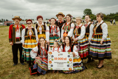 Zdjęcie przedstawia grupę ludzi ubranych w tradycyjne stroje ludowe. Trzymają dużą kartę z napisem: "Nagroda za zajęcia drugiego miejsca w finale dziesiątej edycji konkursu kulinarnego pn. Bitwa Regionów. Piętnaście tysięcy złotych."