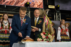 Marszałek Jarosław Stawiarski oraz Starosta  Waldemar Jakson podczas obrzędu krojenia chleba na scenie dożynkowej. Za nimi widoczne młode dziewczęta przyodziane w stylu ludowym.