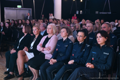 Zdjęcie przedstawiające publikę podczas wydarzenia. Miejsca siedzące na przyciemnionej sali są kompletnie wypełnione. Wśród widowni widoczni są liczni policjanci oraz policjantki.