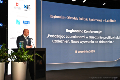Marszałek Województwa Lubelskiego Jarosław Stawiarski na mównicy przemawiający do publiczności. Za nim, na ekranie, wyświetlony jest napis: "Regionalna Konferencja: 'Podążając za zmianami w dziedzinie profilaktyki uzależnień. Nowe wyzwania do działania.'"