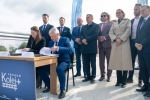 Około dziesięciu osób, wiekszość stoi. Na pierwszym planie stoi ustawiony stolik przy którym siedzą kobieta i mężczyzna i podpisują umowę.