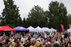 Zdjęcie przedstawiające tłum siedzących ludzi wpatrzonych wspólnie w jeden punkt poza kadrem. Za nimi widoczny jest rządek stoisk.