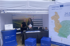 Zdjęcie przedstawiające stoisko Departamentu Strategii i Rozwoju Urzędu Marszałkowskiego Województwa Lubelskiego. Nad dwoma kobietami pozującymi do zdjęcia widoczny jest szyld z napisem: "Regionalny Punkt Kontaktowy Programu Interreg Next Polska - Ukraina 2021-2027 w Lublinie". Bezpośrednio przed stoiskiem widoczna jest ścianka z mapą przedstawiającą regiony uczestniczące w projekcie. 