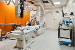 Na zdjęciu widoczne jest szpitalne pomieszczenie z maszynami. W centrum pokoju stoi duża, podłużna maszyna z licznymi kablami oraz monitorami. Za nią, ściana, wyłożona jest serią szafek po całej długości.