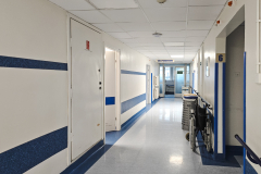 Zdjęcie przedstawiające szpitalny korytarz. Biały i sterylny, ozdobiony niebieskimi paskami biegnącymi wzdłuż jego długości. Oprócz kilku par drzwi ścielących ściany korytarza można zauważyć, między innymi, złożony wózek inwalidzki postawiony przy rozwidleniu korytarza.
