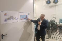 Mężczyzna z mikrofonem przemawia wskazując na jeden z dwóch obrazków zawieszonych na szerokich drzwiach. Obrazki przedstawiają budowę mechaniczną helikopterów.