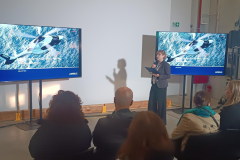 Kobieta z mikrofonem przemawia do publiczności. Po jej obu stronach znajdują się ekrany przedstawiające helikoptery.