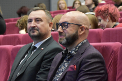 Zbliżenie na dwóch siedzących na sali wykładowej mężczyzn. W tle zarysy innych siedzących osób. 