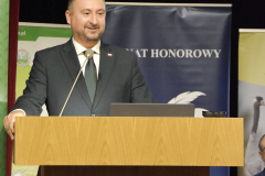 Człowiek przy mównicy przed mikrofonem. W tle banery partnerów projektu. Pod mównicą ekran z planszą informacyjną o  Wielkiej Lekcji "Człowiek w środowisku".