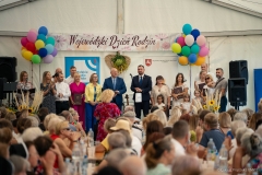 Marszałek Jarosław Stawiarski stoi na scenie i zwraca się do zebranych. Widać około kilkudziesięciu osób siedzących na publiczności. Scena znajduje się pod dużym namiotem i jest ozdobiona bukietami z kwiatów i zbóż