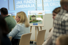 Zdjęcie przedstawia część widowni podczas wydarzenia. Obok widowni rozstawione są sztalugi z obrazami.