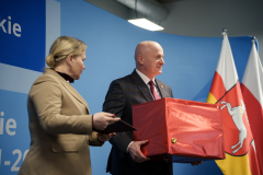 Marszałek Województwa Lubelskiego Jarosław Stawiarski oraz Dyrektor Departamentu Zarządzania Programami Regionalnymi Anna Brzyska przemawiający na scenie. MArszałek trzyma duże czerwone pudło. Za nimi widoczne są flagi Województwa Lubelskiego oraz Polski.