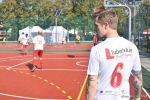 Mecz w ramach akcji "z podwórka na boisko". Zawodnik drużyny urzędników w białej koszulce, stoi plecami do obiektywu. Na koszulce logo lubelskie smakuj życie. poniżej czerwona cyfra sześć. w tle rozgrzewający się na bosiku zawodnicy urzędu.