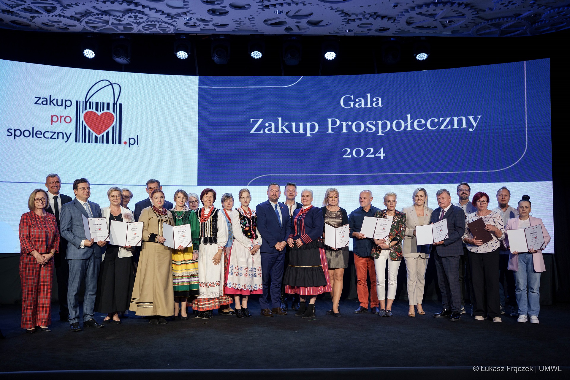 Wicemarszałek Marek Wojciechowski stoi na scenie i wręcza kobiecie certyfikat, za nimi stoi około dziesięciu innych laureatów