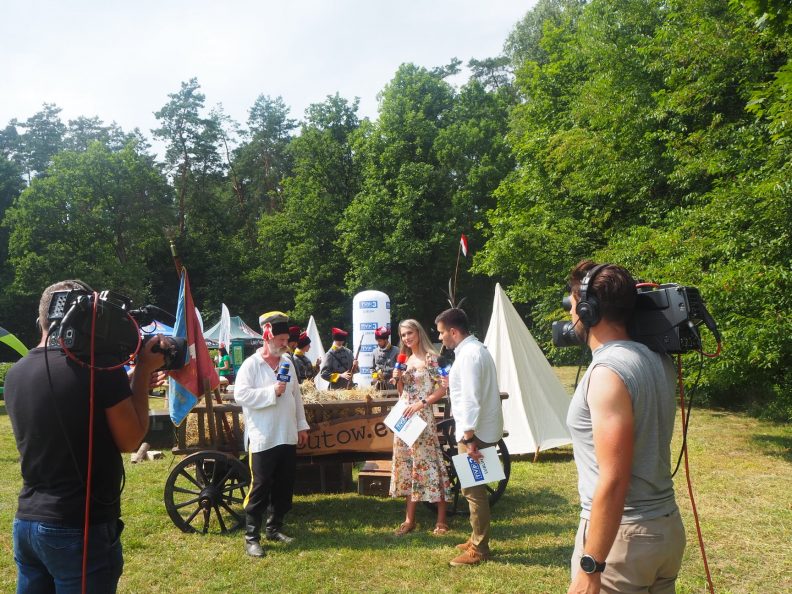 Zdjecie przedstawiające mężcyznę w stroju ludowych który stojąc przy drabiniastym wozie udziela wywiadu dwójce reporterów. Na pierwszym planie stoi kamerzysta nagrywający cały wywiad.