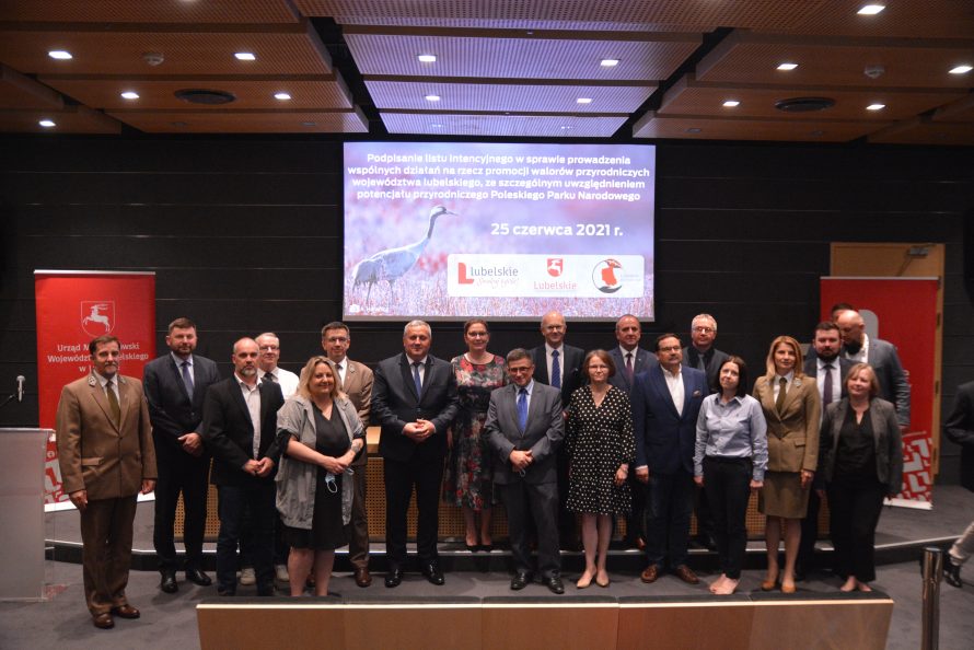 Zdjęcie ukazujące grupę ludzi stojacą w sali konferencyjnej. Za nimi znajduje się ekran z wyświetlaną prezentacją nt. żurawi