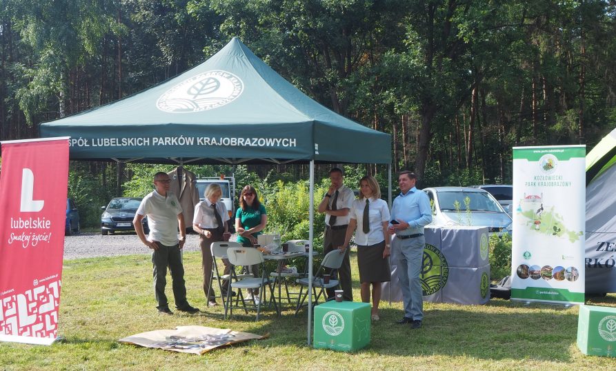 Zdjęcie przedstawiające rozłożony namiot a pod nim grupę stojących ludzi, zebranych wokół stołu z krzesłami