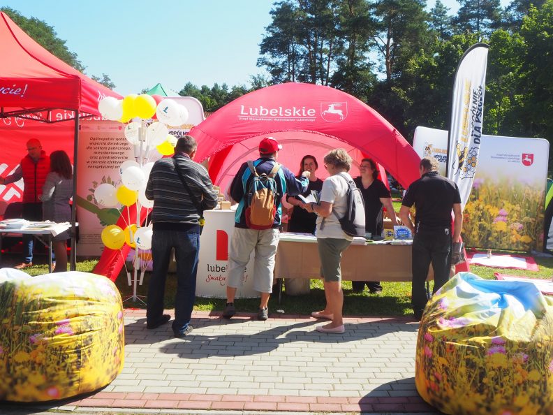 Zdjęcie ukazujące rozłozony namiot festynowy w którym dwie kobiety prezentuja foldery czterosobowej grupie osób