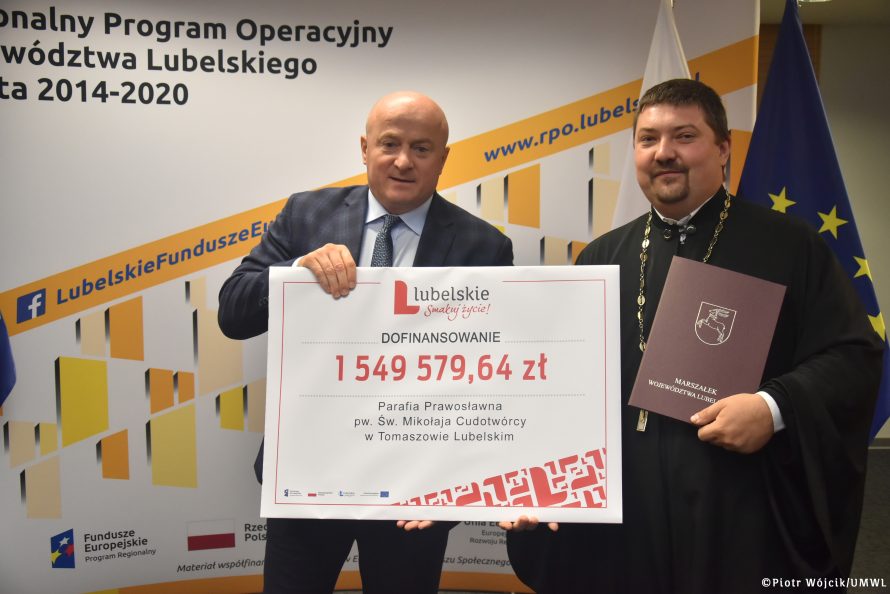 Wręczenie umów na dofinansowania remontów i konserwacji zabytkowych obiektów sakralnych. Od lewej marszałek Jarosław Stawiarski, ks. Dariusz Wasiluk. Obaj trzymają symboliczny czek. Na czeku od góry logo lubelskie smakuj życie, poniżej napis czarną czcionką dofinansowanie, poniżej czerwoną cziocnką kwtoa 1549579,64 zł, poniżej napis parafia prawosławna pw. Św. Mikołaja Cudotwórcy w Tomaszowie Lubelskim