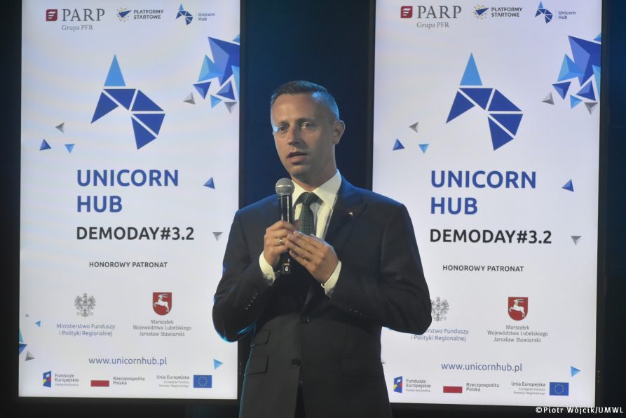 Wicemarszałek Województwa Lubelskiego Michał Mulawa w trakcie przemowy. W tle dwa ekrany wyświetlające grafikę wydarzenia Unicorn Hub DemoDay #3.2