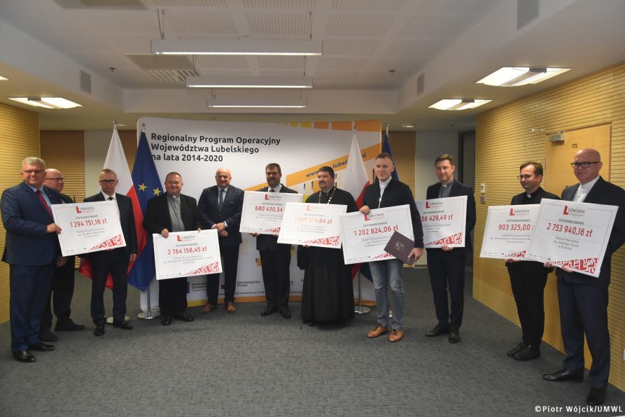 Wręczenie umów na dofinansowania remontów i konserwacji zabytkowych obiektów sakralnych. Zdjęcie grupowe beneficjentów. W rękach trzymają symboliczne czeki z wypisanymi kwotami dofinansowania.