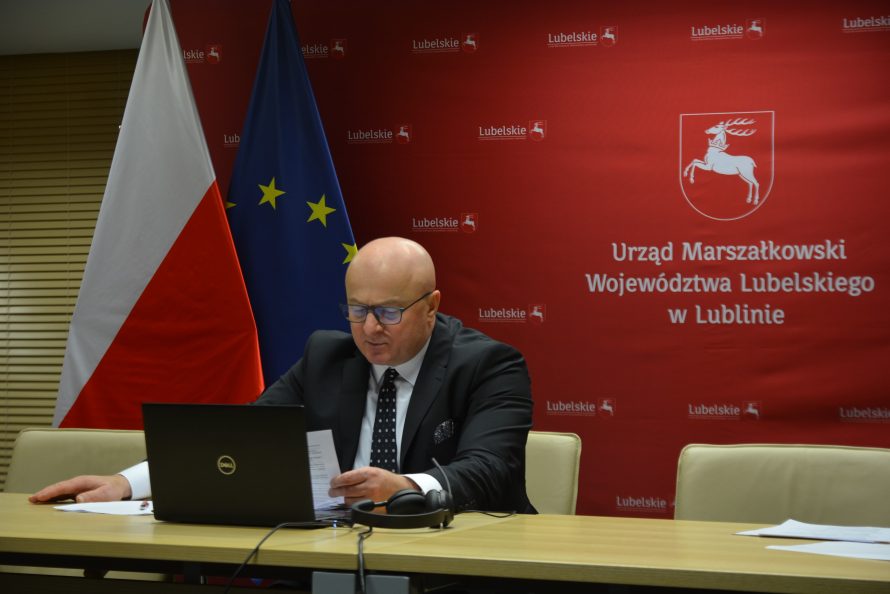 Marszałek Stawiarski siedzi przed komputerem, trzymając kartkę z przemówieniem. w tle czerwona ścianka z herbem województwa i napisem urząd marszałkowski województwa lubelskiego. po lewej od marszałka stoją flagi polski i unii europejskiej
