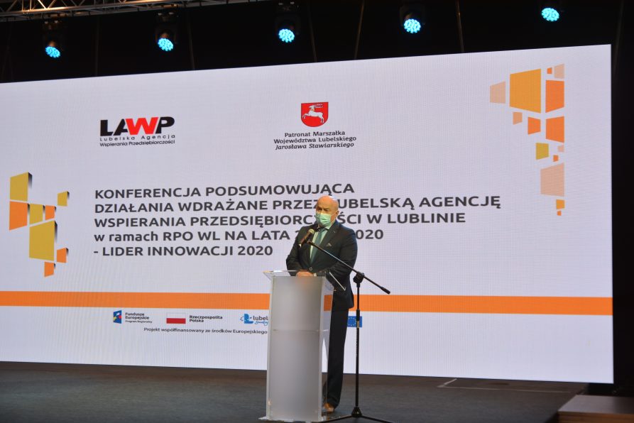 Marszałek Stawiarski w trakcie wideokonferencji stoi przy mównicy. w tle ekran wyświetlający napis konferrencja podsumowująca działania wdrażane przez LAWP