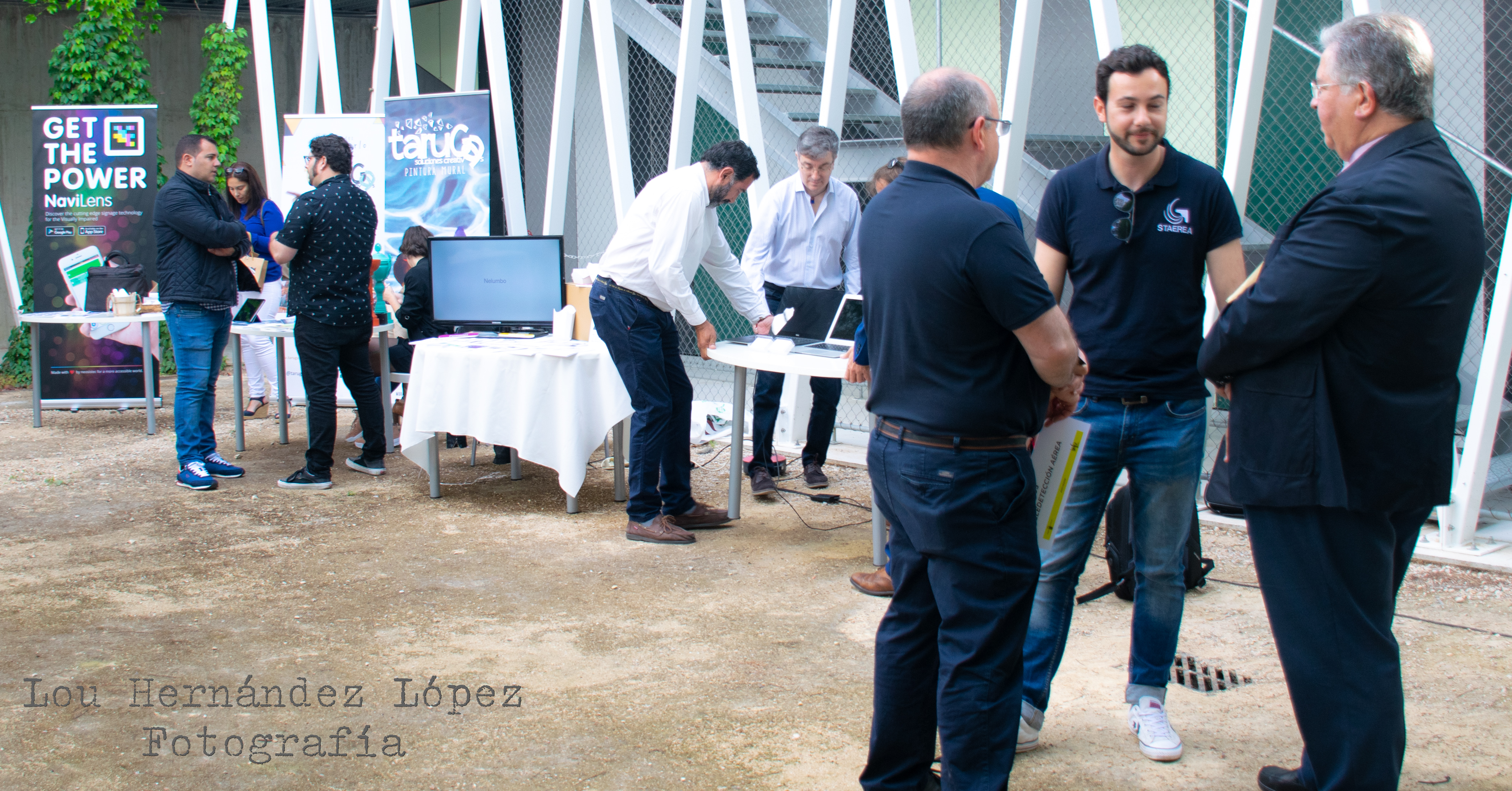 Z prawej grupa trzech rozmawiających mężczyzn, w tle inne postacie w trakcie rozmowy, przy stole.