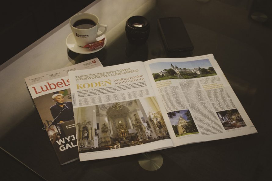 Dwa egzemplarze gazety lubelskie, jeden rozłożony przykrywa okładkę drugiego, leżą na szklanym blacie w towarzystwie telefonu, obiektywu, i filiżanki z kawą