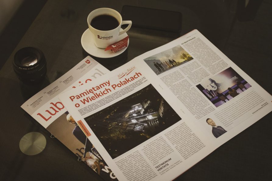Dwa egzemplarze gazety lubelskie, jeden rozłożony przykrywa okładkę drugiego, leżą na szklanym blacie w towarzystwie telefonu, obiektywu, i filiżanki z kawą