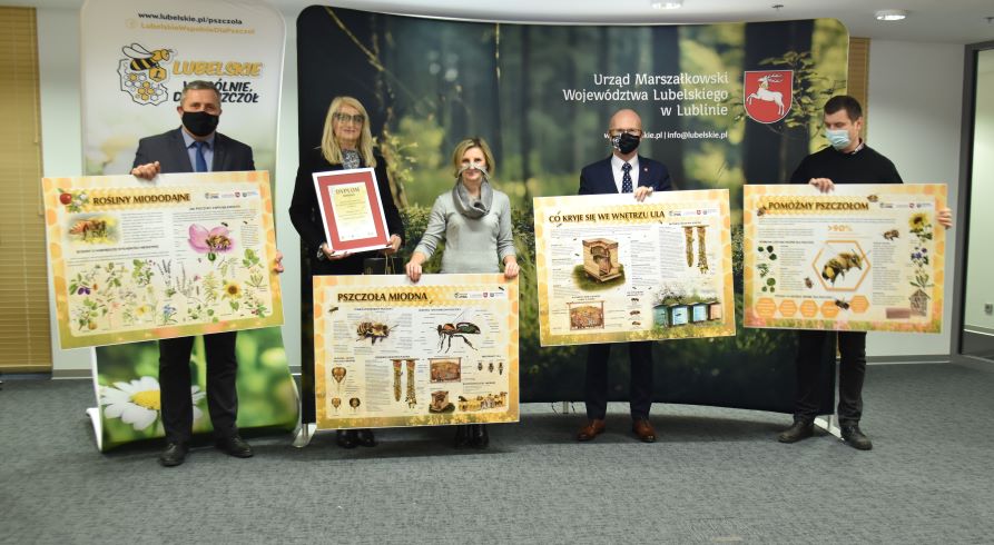 Na zdjęciu widać pięciu uczestników spotkania (reprezentantów organizatorów konkursu i dyrektor nagrodzonej szkoły), którzy trzymają w rękach duże tablice edukacyjne o tematyce pszczelej. W tle za nimi widać ściankę z przyrodniczą fotografią i napisem „Urząd Marszałkowski Województwa Lubelskiego” oraz stand z logo kampanii „Lubelskie – wspólnie dla pszczół”