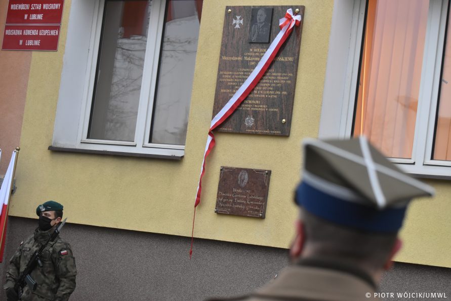 Tablica pamiątkowa przepasana biało-czerwoną szarfą. W dolnym lewym rogu żołnierz warty honorowej