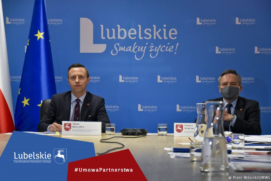 Na zdjęciu przy stole siedzą od lewej wicemarszałek Michał Mulawa i wicemarszałek Zbigniew Wojciechowski. w tle niebieska ścianka z logo lubelskie smakuj życie. Po lewej flaga Polski i Unii Europejskiej. Na dole na niebieskiej geometrycznej apli logotyp lubelskie, obok, na czerwonej geometrycznej apli napis #UmowaPartnerstwa
