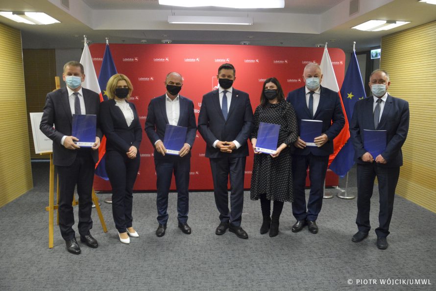 Na zdjęciu w szeregu stoi 7 osób, część z nich trzyma teczki z umowami, po środku stoi członek zarządu województwa Zdzisław Szwed