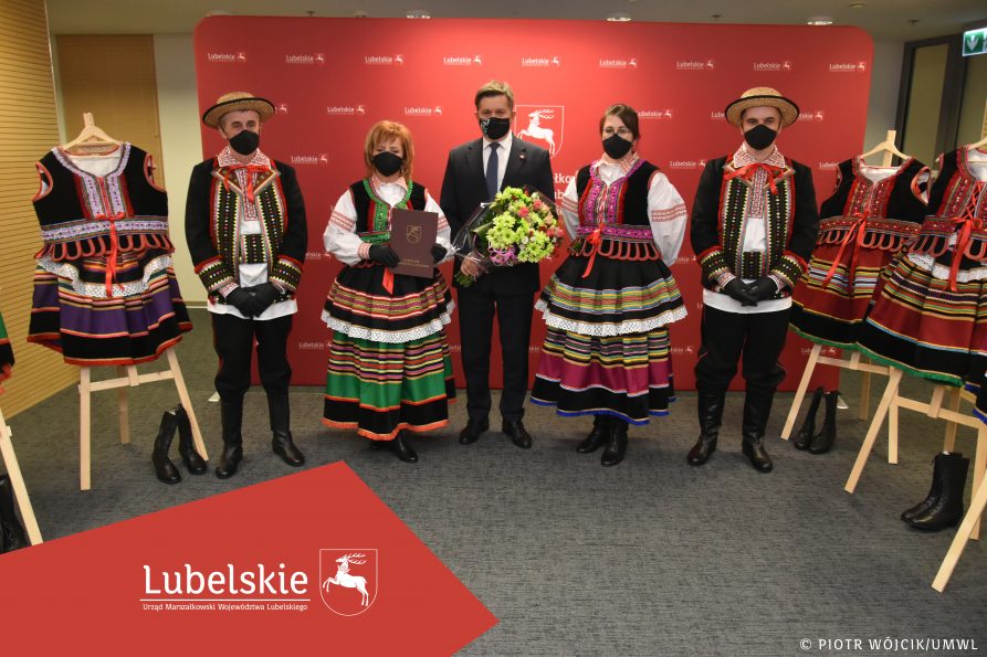 Grupowe zdjęcie - po środku stoi członek zarządu Zdzisław Szwed. Po obydwu stronach stoją członkowie Koła Gospodyń Wiejskich. Po bbokach kadru na sztalugach rozwieszone stroje ludowe.