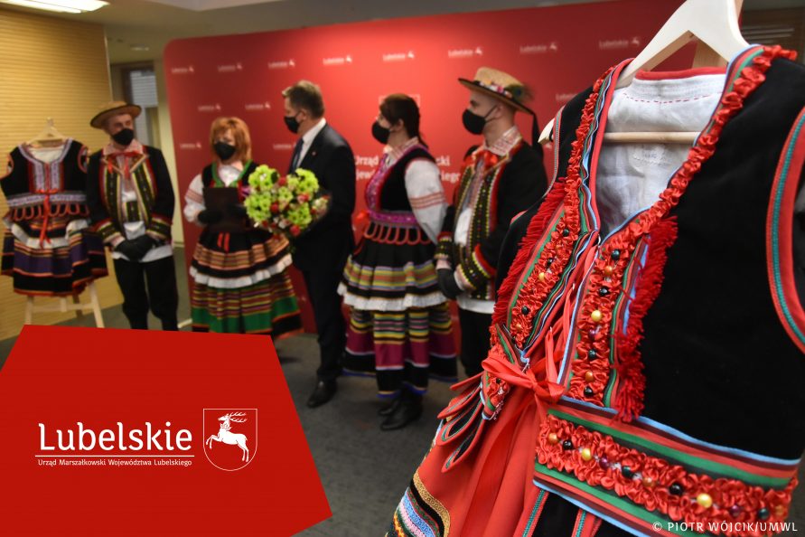 Na pierwszym planie z prawej jeden ze strojów ludowych. po lewej na geometrycznej czerwonej apli logotyp lubelskie. W tle, lekko rozmyty obraz, członek zarządu Zdzisław Szwed wśród członków jednego ze zwycięskich Kół.