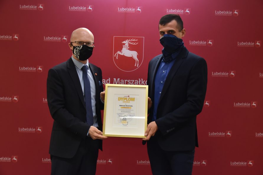 Na zdjęciu od lewej stoją Członek Zarządu Województwa Lubelskiego, Sebastian Trojak oraz Mateusz Skowronek. Panowie wspólnie trzymają dyplom uznania za zdjęcie stycznia na tle ścianki fotograficznej z logotypami i herbem Urzędu Marszałkowskiego Województwa Lubelskiego