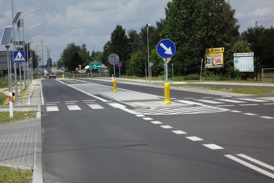 Widok na fragment drogi wojewódzkiej nr 812. Jezdnia z namalowanymi pasami, wysepką przez którą przeprowadzone jest przejście drogowe. W tle ciąg drzew. po obu stronach drogi widoczne są latarnie oraz znaki drogowe.